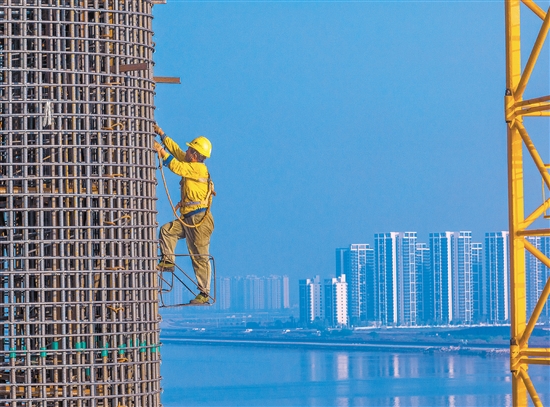 奋战在百米索塔之上的建设者身影，与远处的高楼大厦遥呼相应。