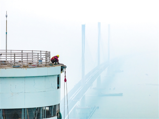 建设者在塔柱上施工，远处“海上长龙”若隐若现。